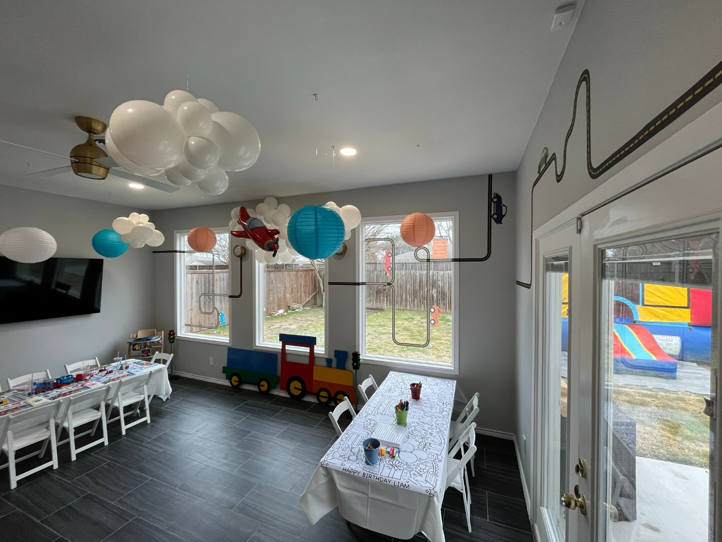 Liam's transportation birthday party room with cloud balloon installation, airplane garland, road decal on walls, coloring tablecloth, and bounce house visible through glass door